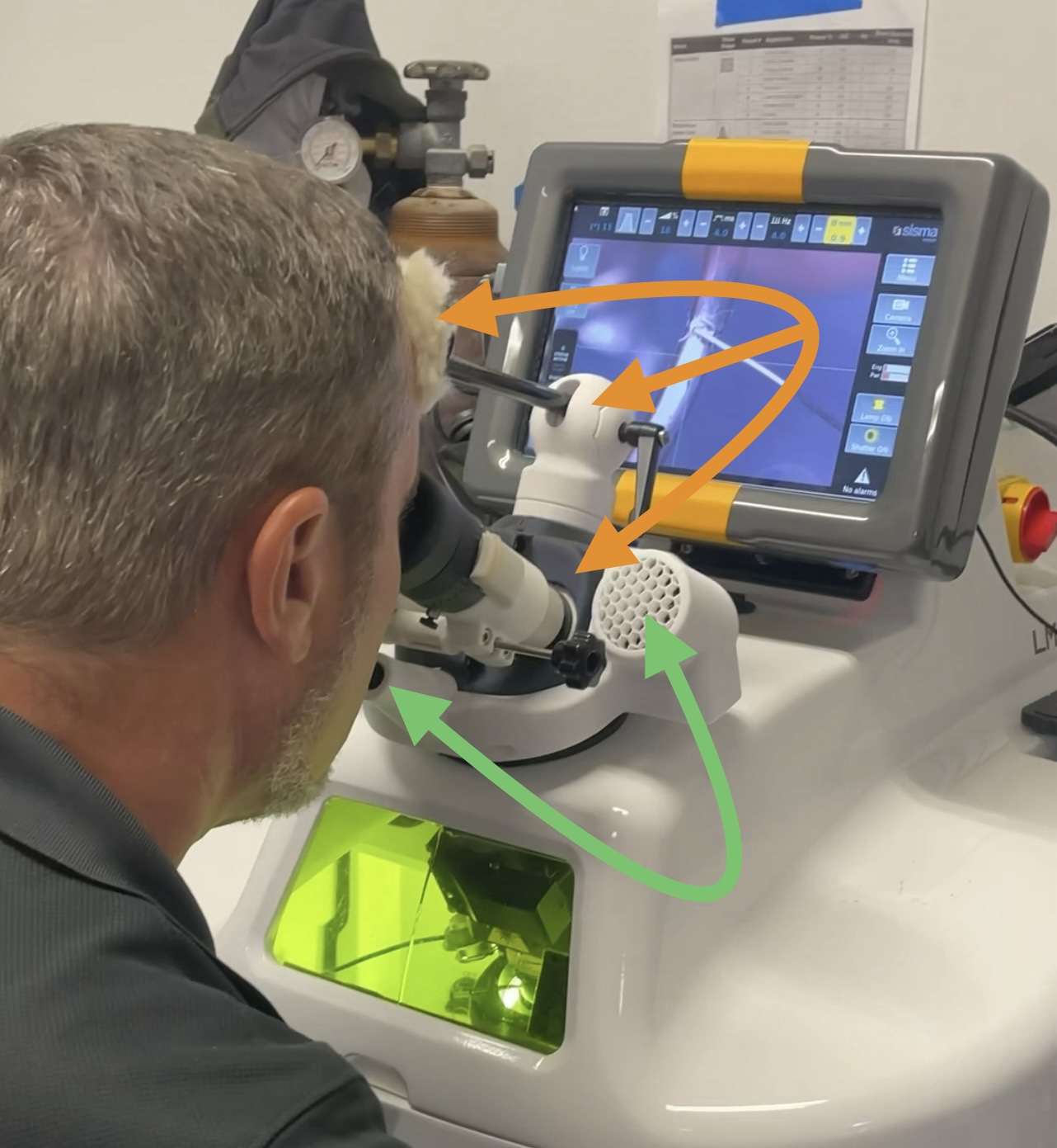 A jeweler looking through the eyepieces of a laser welder with a Leica 1 headrest and the Mic Speaker Assembly fitted. Orange arrows mark the headrest; green arrows mark the MSA microphone and speaker.